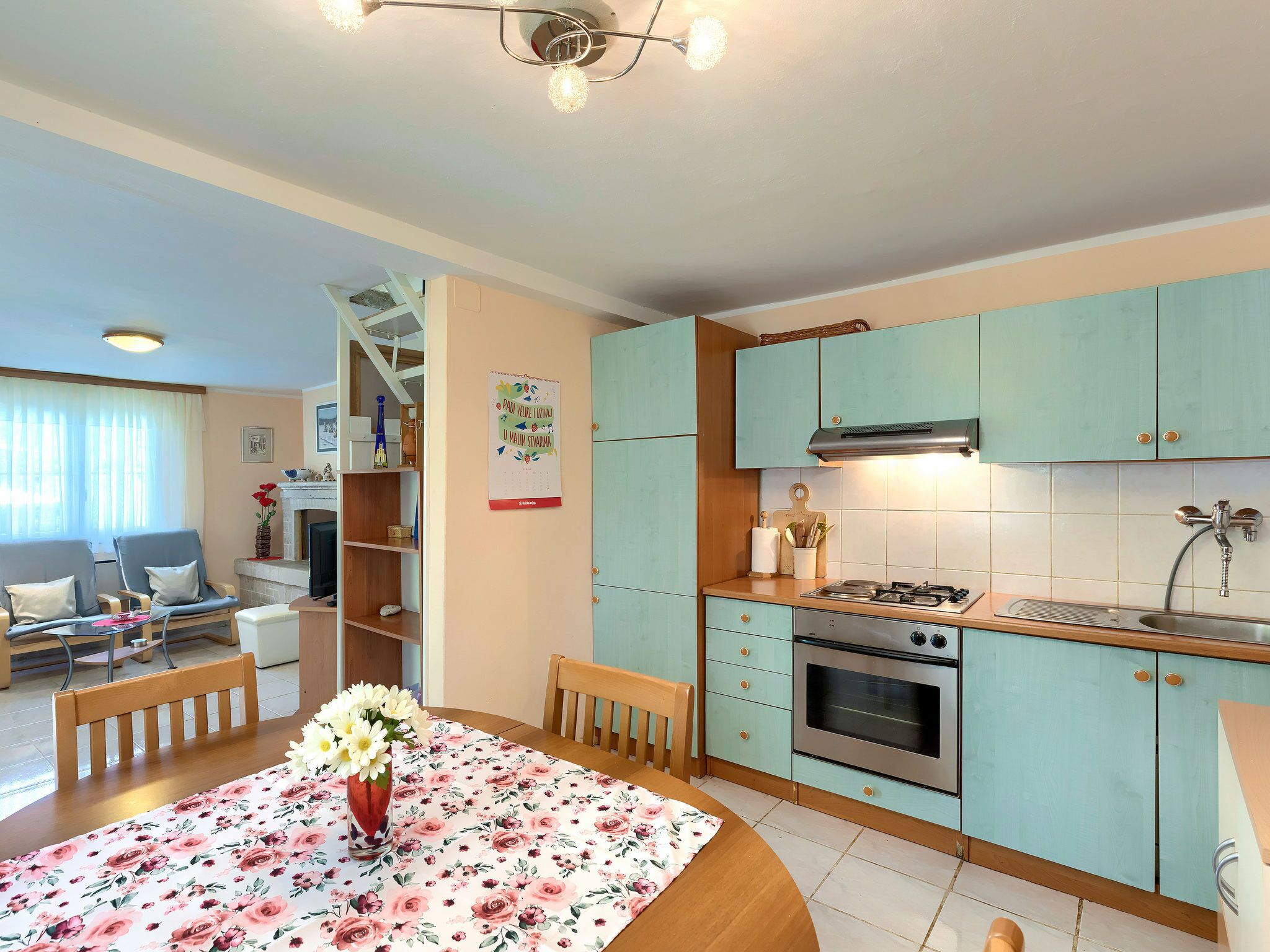 Una cocina con armarios color turquesa, una estufa a gas y un lavamanos. Hay una mesa de comedor con mantel floral y un jarrón de flores.