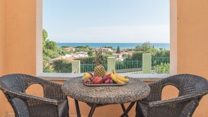Varanda com cadeiras de vime e uma mesa com frutas. Uma vista do mar e edifícios é visível através de um arco.