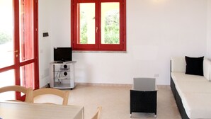 Quarto com mesa de jantar, cadeiras, sofá, uma pequena televisão em um suporte e uma janela com molduras vermelhas.