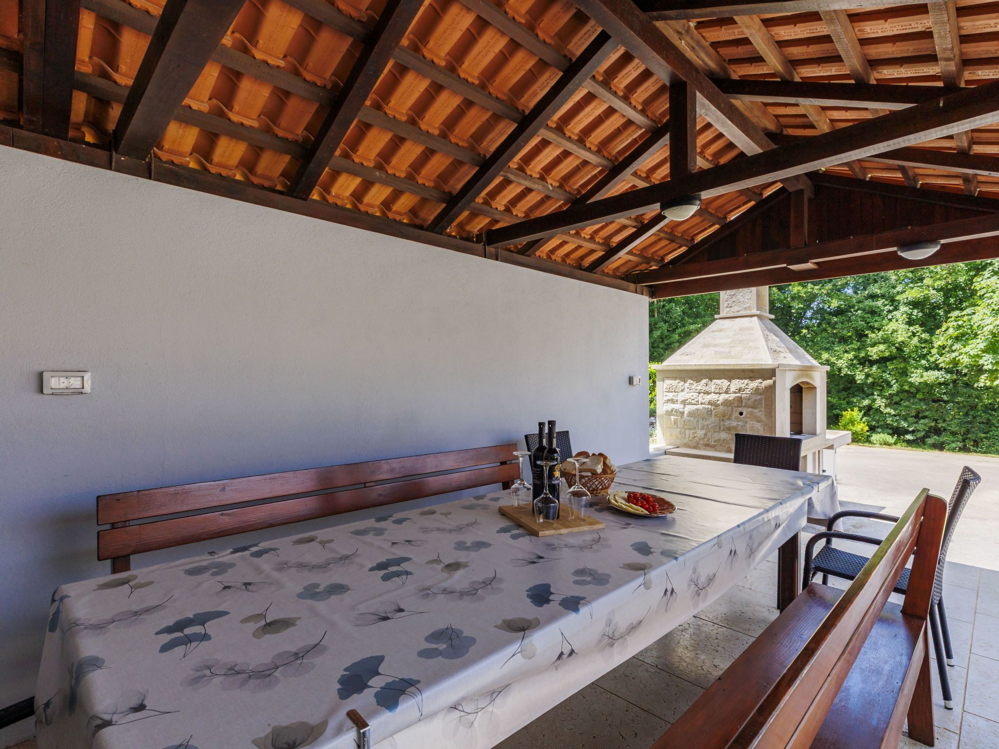 An outdoor dining area with a table set for two, a stone oven in the background, and a bench to the left.