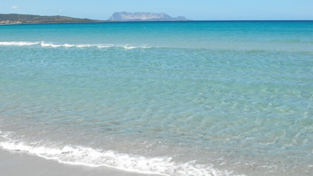 Una playa con agua turquesa cristalina, olas suaves y una cadena montañosa distante.