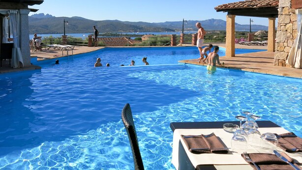 Una piscina amb persones nedant, una taula preparada per dinar i una vista de muntanyes al fons.
