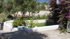 Un edifici modern amb façana blanca, un arbre gran al primer pla i una vista del mar i les colines al fons.