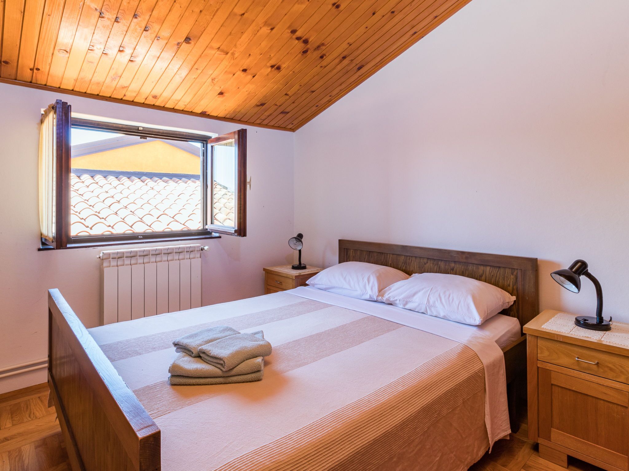 Une chambre avec un plafond en bois, une fenêtre donnant sur un toit recouvert de tuiles, un lit avec du linge de maison blanc, deux tables de chevet avec des lampes et un radiateur.