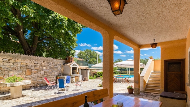 An outdoor patio area with a wooden table, a bottle of wine, and a glass. There are stone walls, a tree, and a pool in the background. - Villa San Giacomo by Interhome (Poreč/Kaštelir)