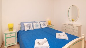 Un dormitorio con colcha azul, almohadas blancas con motivos de árboles, una mesita de noche con lámpara, un aparador con cajones y un espejo redondo en la pared.