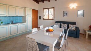 A modern kitchen with blue cabinets, a dining area with a white table and patterned chairs, a blue sofa, and a wooden ceiling.