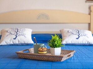 A bed with a blue bedspread, two pillows with a tree design, a tray with a plant, a mug, and a basket.