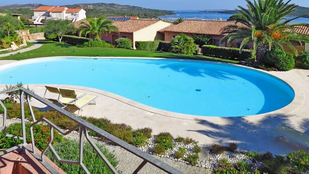 Ein großer Pool mit geschwungener Kante, umgeben von einer angelegten Grünfläche mit Palmen und Blick auf das Meer.