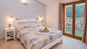 Ein Schlafzimmer mit Bett, Nachttischen, einer kleinen Pflanze und Blick ins Freie durch eine Glastür.