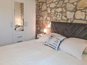 Un dormitorio con pared de piedra, un armario blanco, una cama con ropa de cama blanca y una mesita de noche con lámpara.