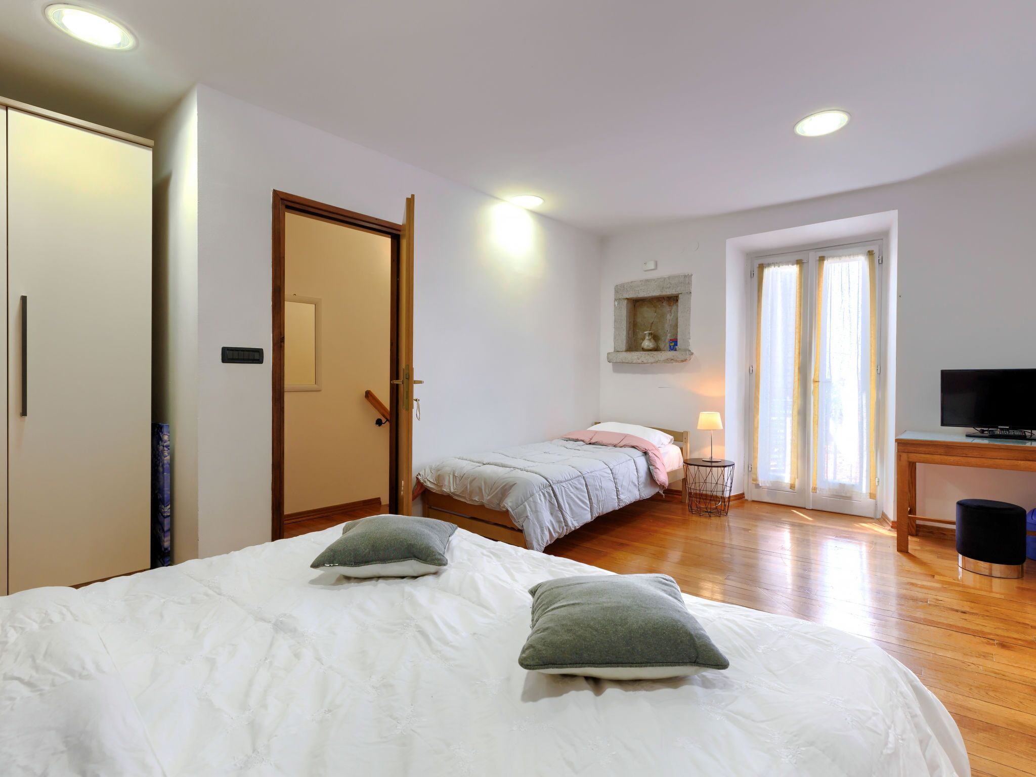 A bedroom with a bed, a desk with a TV, a window with curtains, and a door.