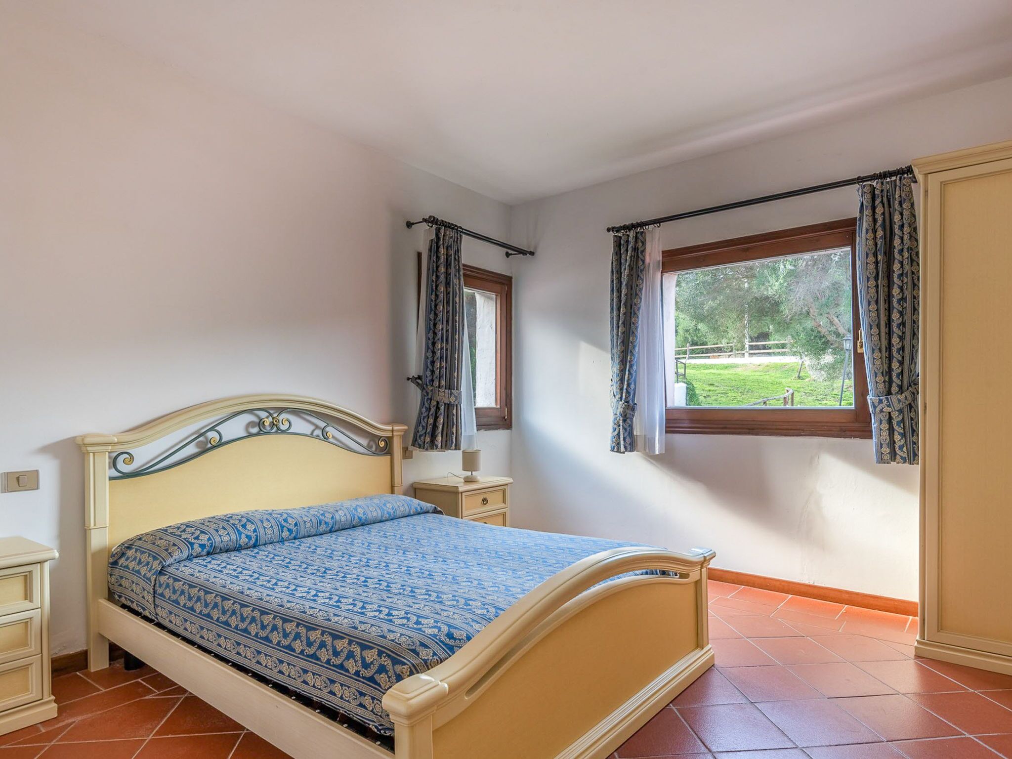 Une chambre à coucher avec un lit, deux tables de chevet, une fenêtre avec des rideaux et une vue sur la verdure.