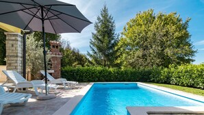 A rectangular swimming pool with a tiled edge, surrounded by a stone deck. There are white lounge chairs and a large umbrella providing shade. - Vacation home Antoni by Interhome (Pićan)