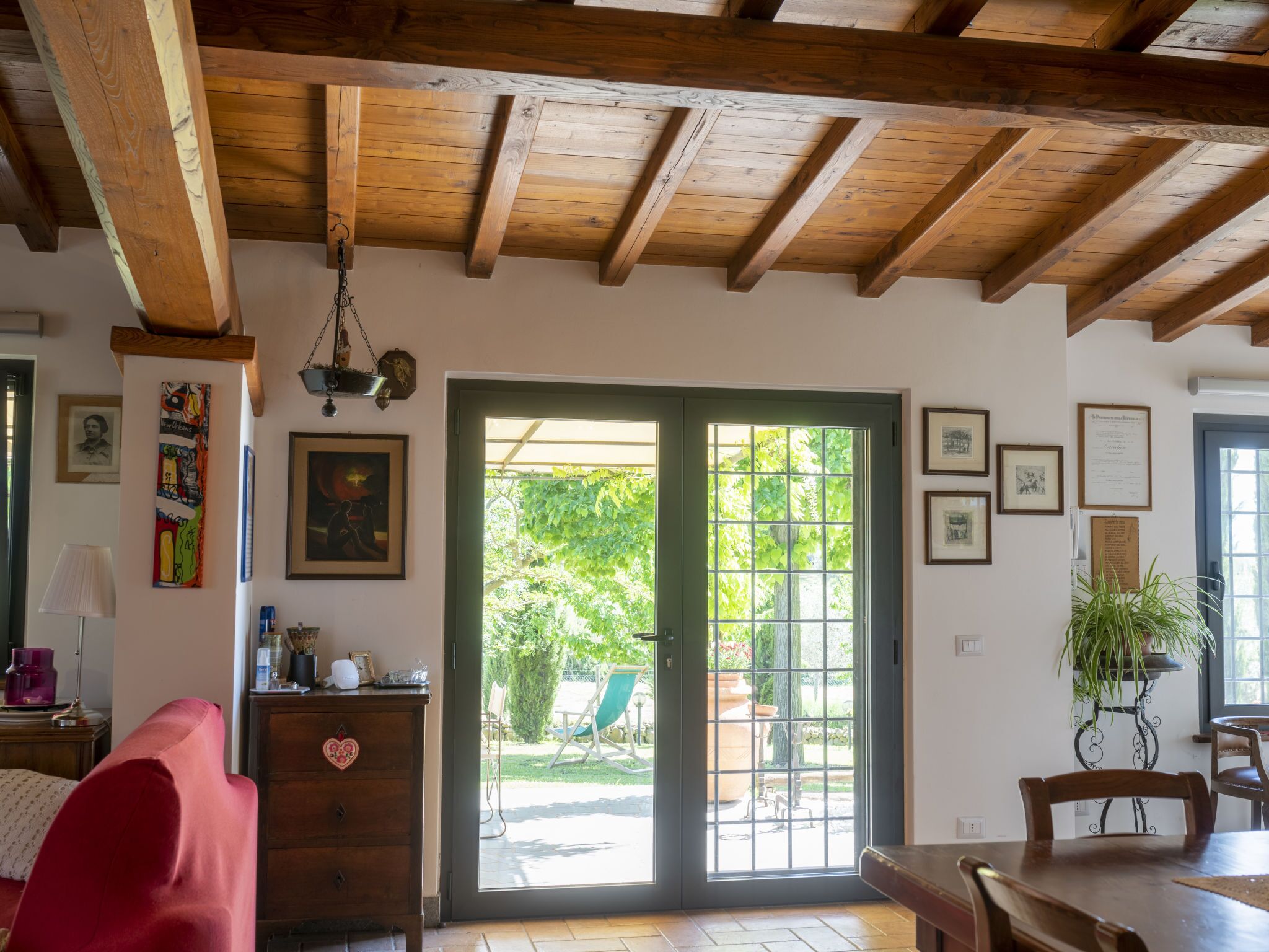 Een kamer met een houten plafond, een schuifdeur van glas naar een tuin, een eettafel en een rode stoel.