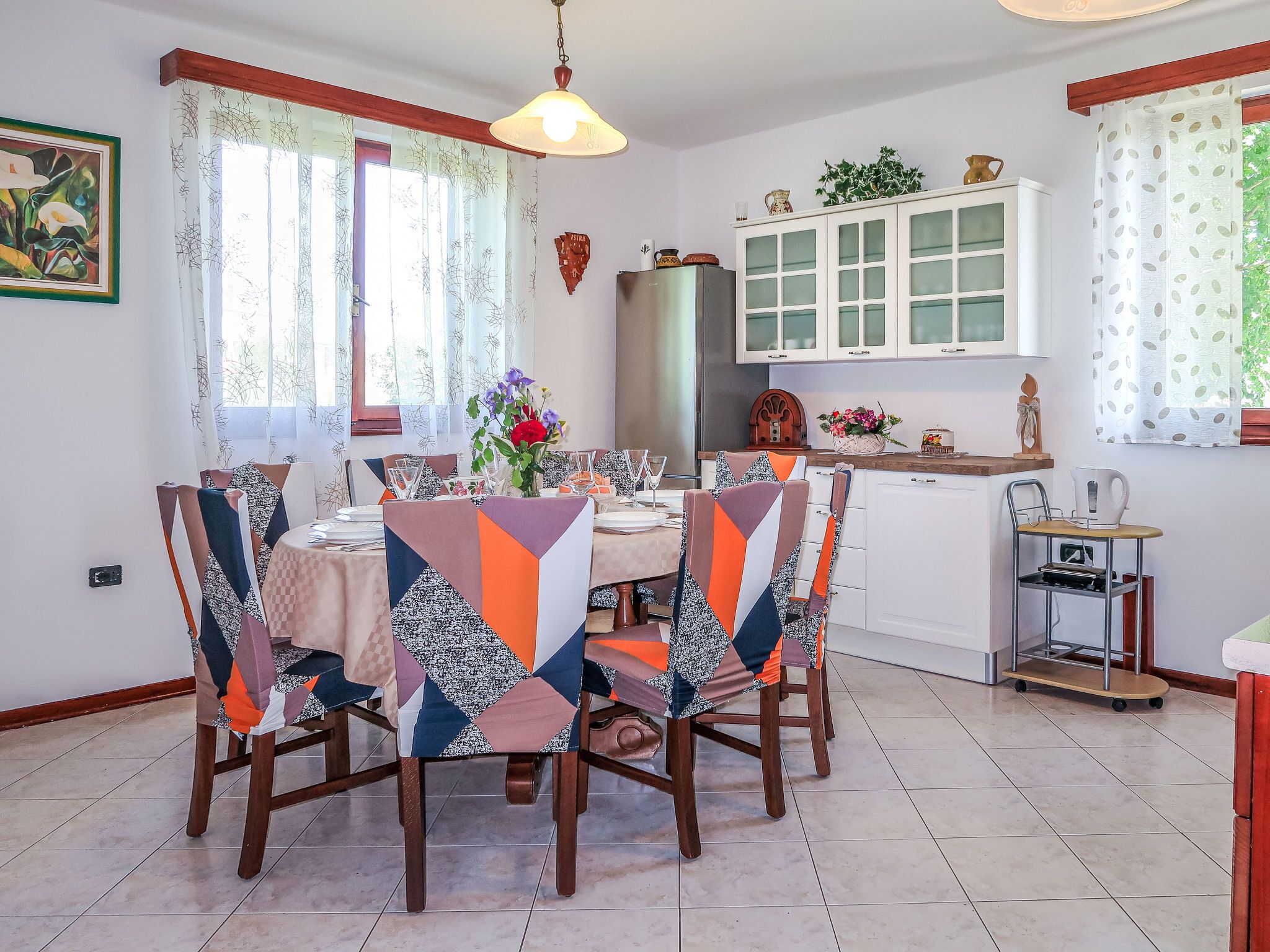 A dining area with a table set for four, surrounded by chairs with patterned upholstery. A decorative vase with flowers is on the table, and a cabinet with glass doors is in the background.