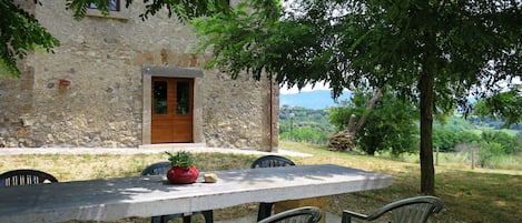 A stone building with a wooden door and windows, a table with chairs, and a view of the countryside.