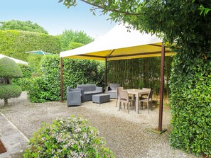 A covered outdoor seating area with a table and chairs, surrounded by greenery and a white canopy.