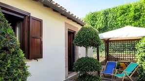 A building with a tiled roof, a door, and a window with shutters. There are two chairs and a table under a covered area.