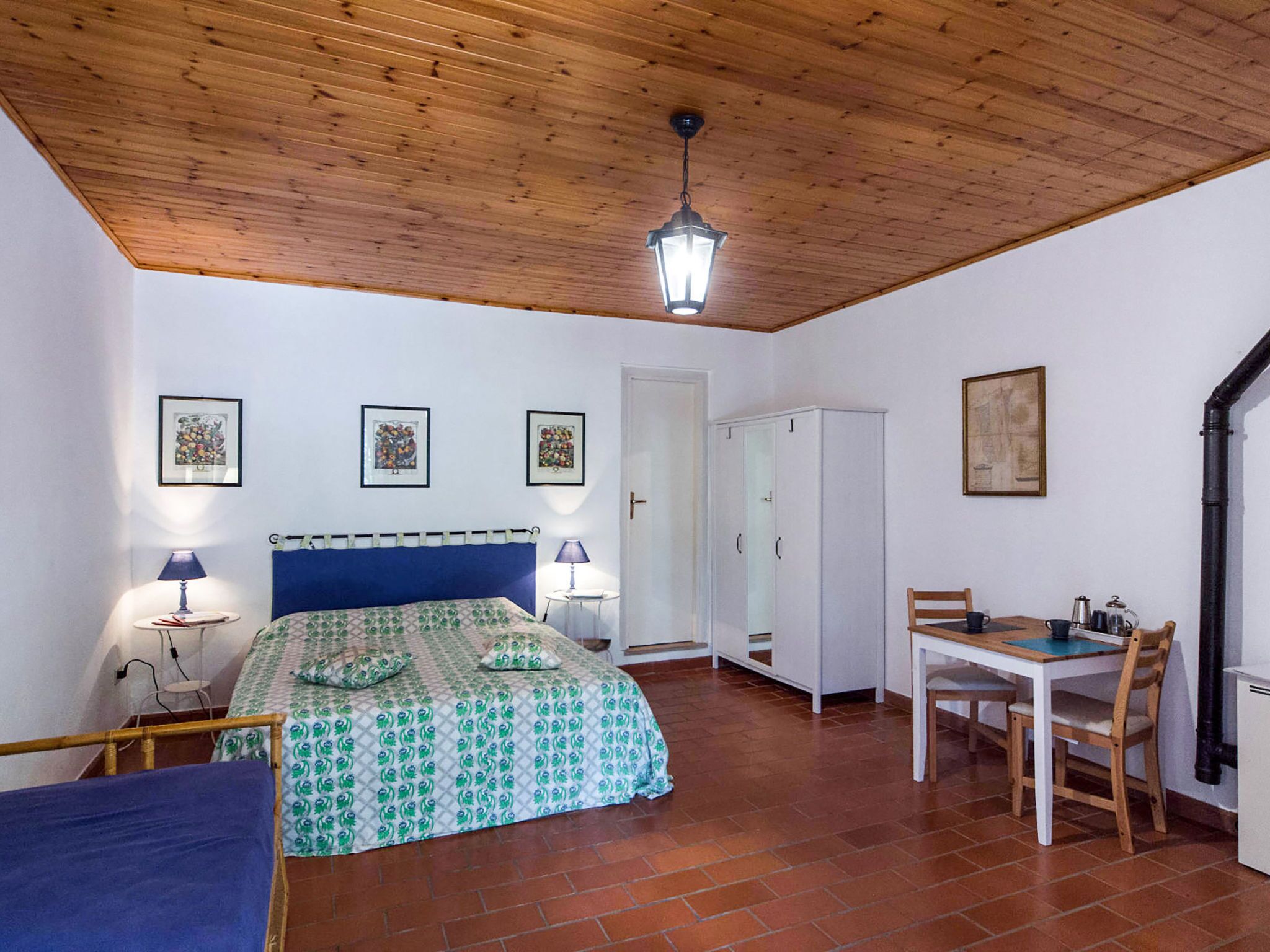 A bedroom with a bed, bedside tables, a wardrobe, a desk with chairs, and a hanging light fixture.