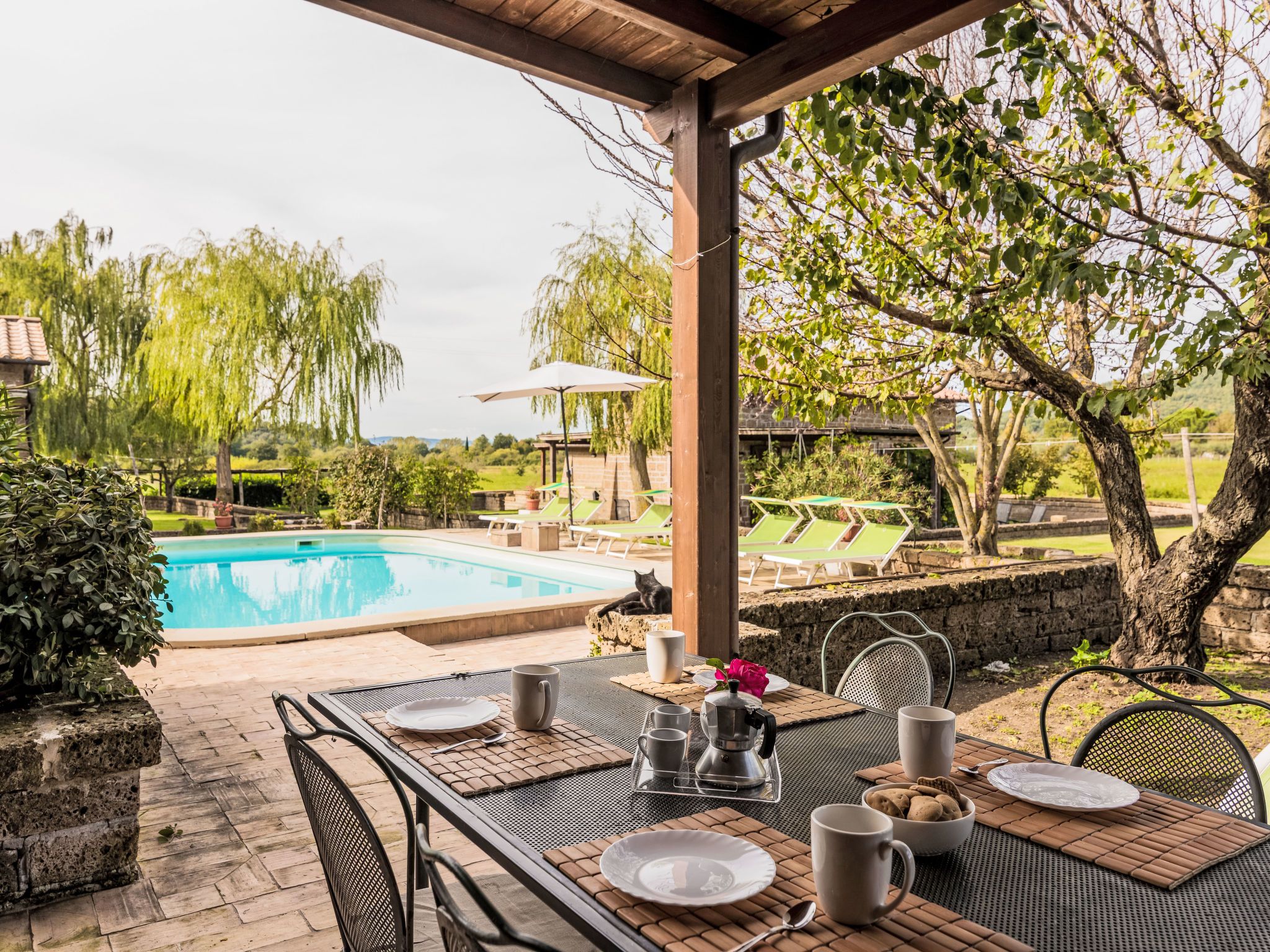 An outdoor dining area with a table set for two, overlooking a swimming pool and a garden with trees and a gazebo.