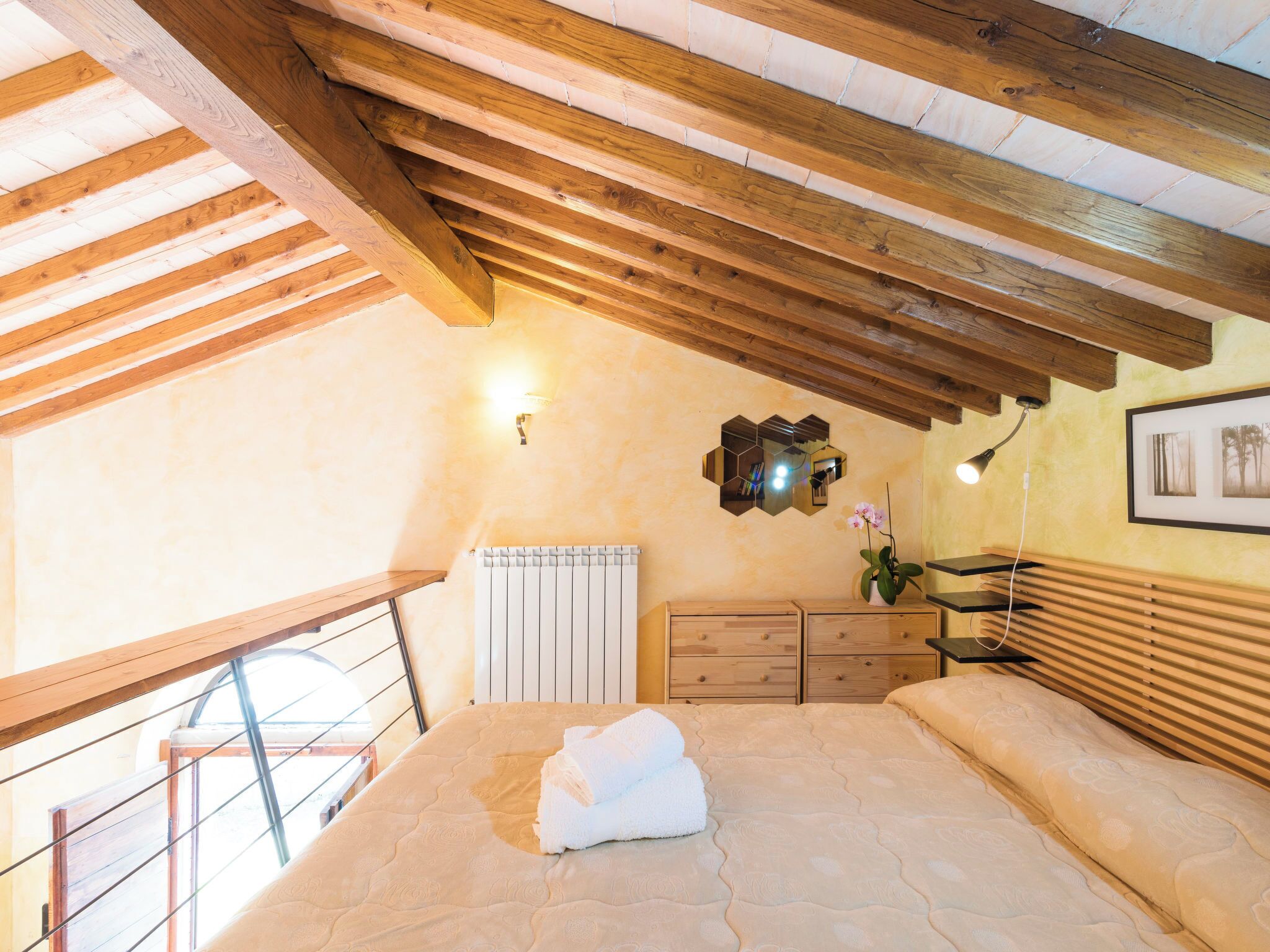 A bedroom with a sloped ceiling, wooden beams, a bed with a mattress and pillows, a dresser, a radiator, a mirror, and a framed picture on the wall.