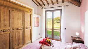 Ein Schlafzimmer mit Holzschrank, einem Bett mit Blumenstrauß, einem Nachttisch und Blick auf einen Garten durch eine Glastür.