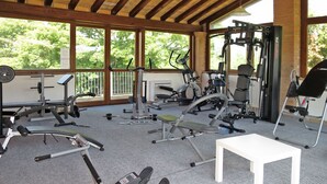 A gym with various exercise equipment, a white bench, and large windows offering a view of greenery.