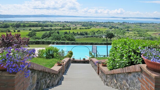 A pool with a stone wall leading to it, surrounded by greenery and a scenic view of fields and hills.