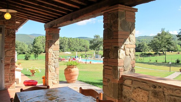 A covered outdoor dining area with a table set for two, a large terracotta pot, and a view of a swimming pool and green landscape. - Alighiero II by Interhome (Boccheggiano)