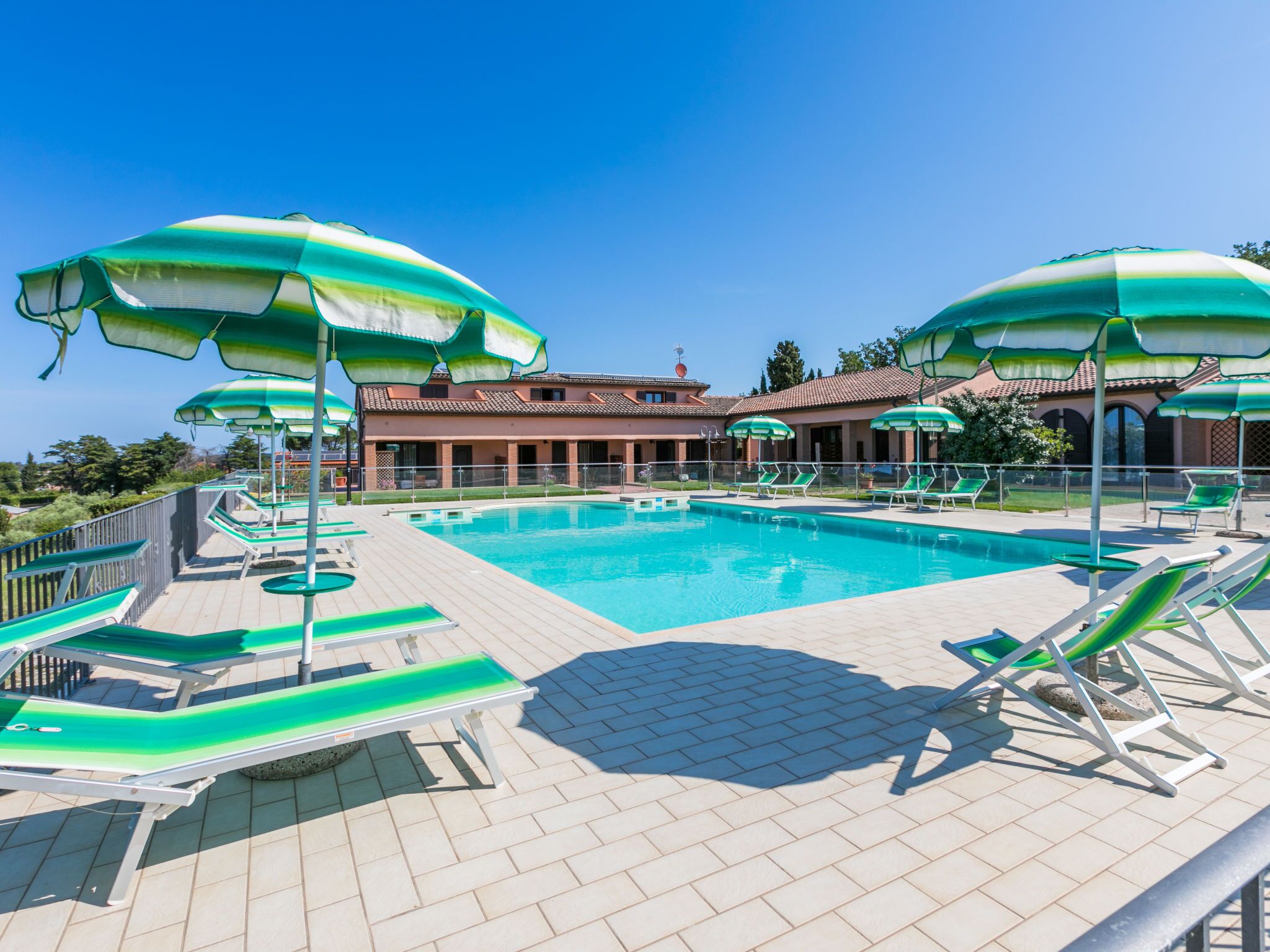 A swimming pool with green and white striped umbrellas, lounge chairs, and a building with a tiled roof in the background.