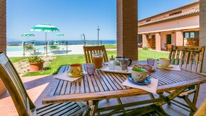 Une table en bois, dressée avec une carafe, des tasses et un verre, donnant sur une piscine et une plage parsemées de parasols.