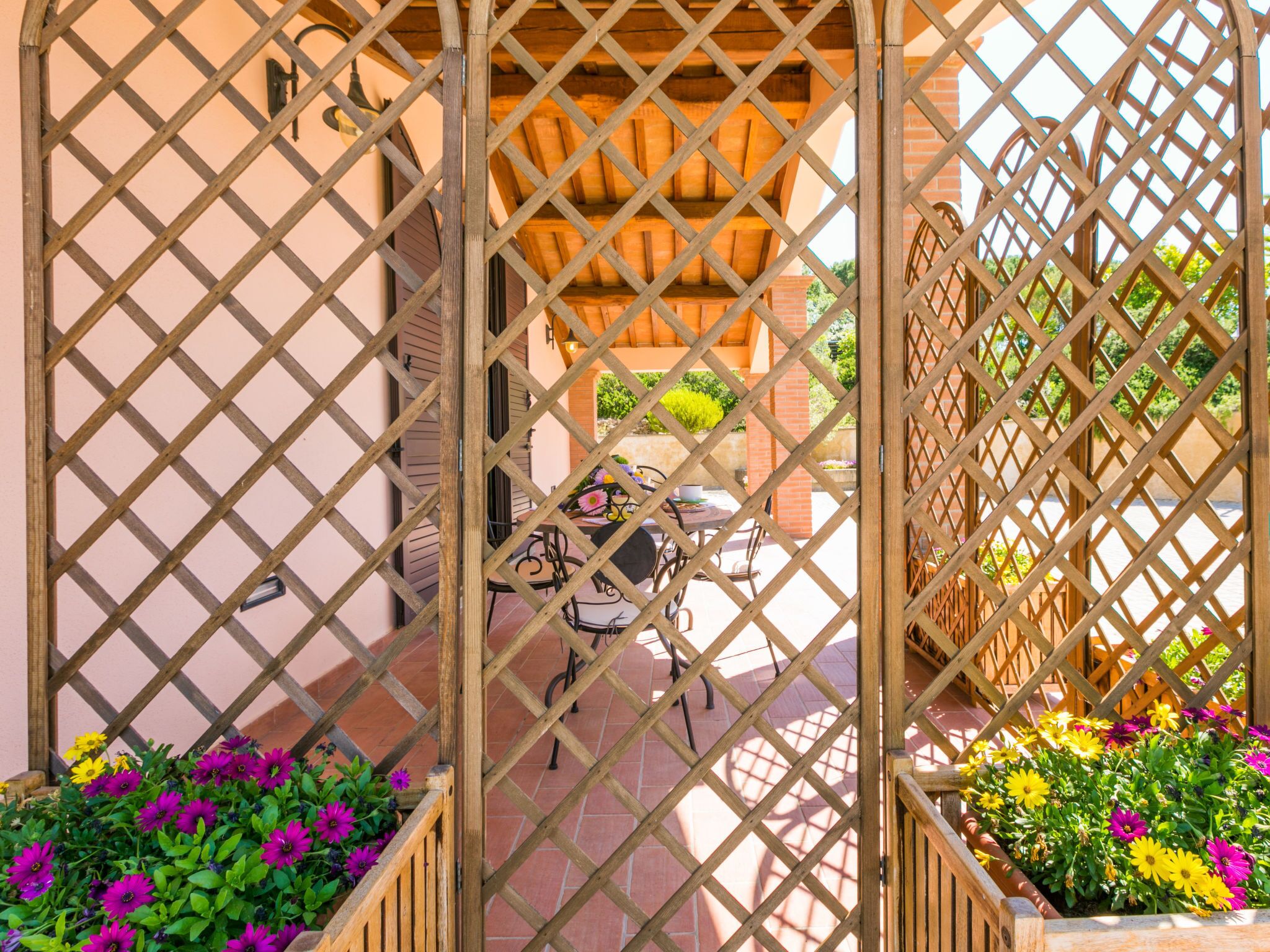 Ein Holzziergitterstor führt zu einer Terrassenanlage mit Tisch und Stühlen, umgeben von Blumenbeeten.