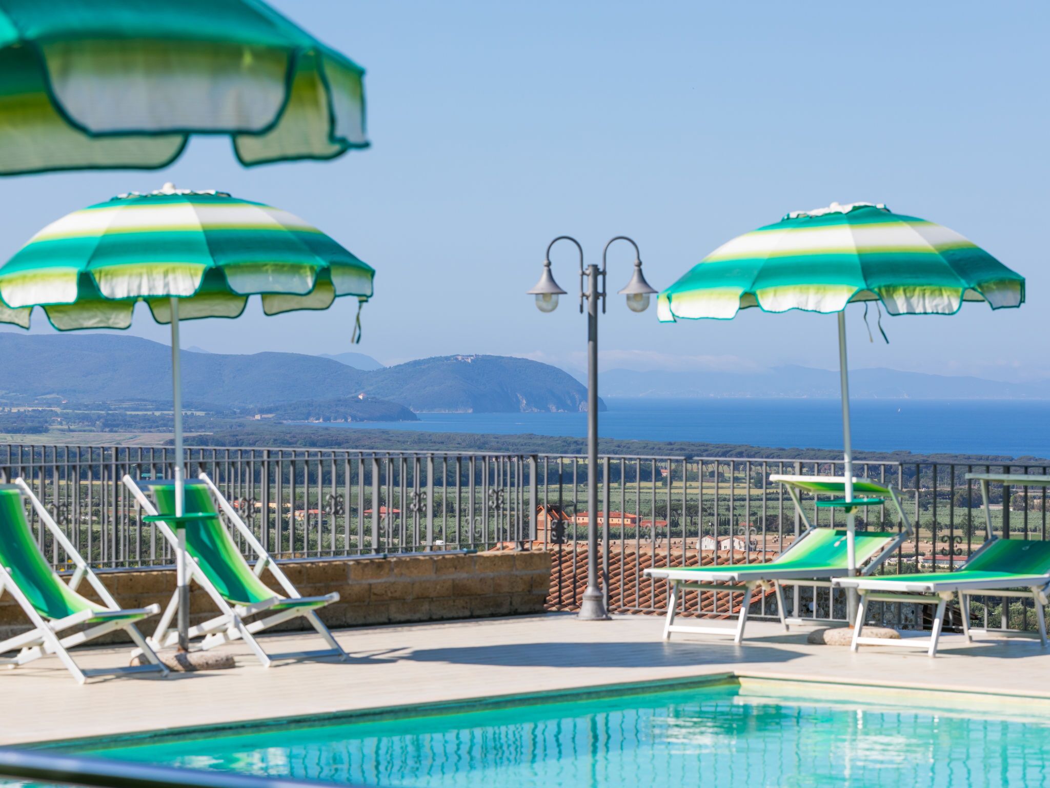 Zona de piscina con sombrillas verde y blanco a rayas, sillas de descanso y vista al mar y montañas de fondo.