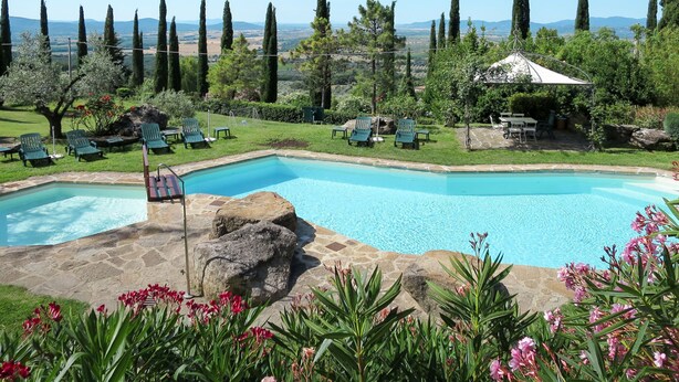 Piscina com espreguiçadeiras e um gazebo, cercada por vegetação e ciprestes.