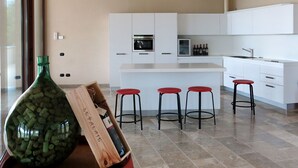 A modern kitchen with white cabinets, a central island with stools, and a wine bottle on a wooden crate. - Le Palaie - 'Mangiatoia' by Interhome (Peccioli)