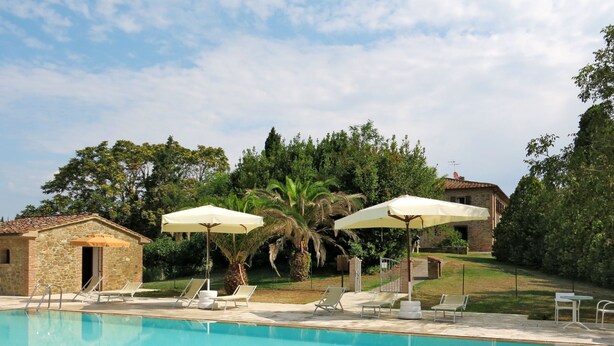 Una piscina con sillas de descanso y sombrillas, rodeada de vegetación y edificios de piedra.