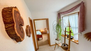 Un pasillo con un cuadro de madera, una planta en maceta, una ventana con cortinas y una cama al fondo.