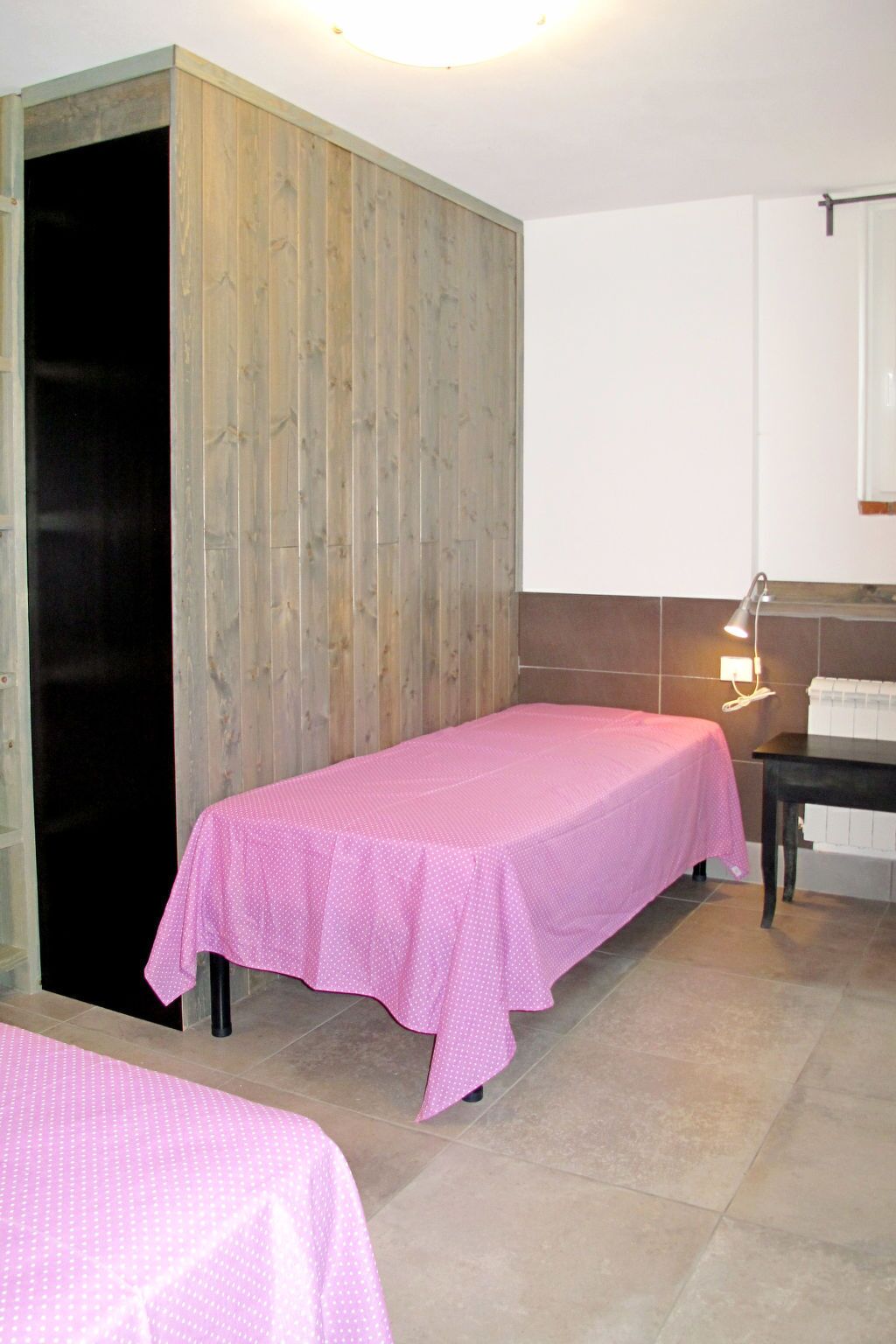 Ein Zimmer mit zwei Betten, Holzvertäfelung an einer Wand, einem kleinen Tisch mit Lampe und einem Heizkörper.