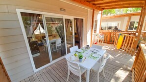 A covered outdoor dining area with a table set for two, sliding glass doors leading to a patio, and a wooden deck with a railing. - Campeggio del Forte by Interhome (Marina di Bibbona)