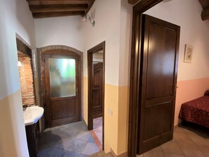 A narrow hallway with a tiled floor, a wooden door, a mirror, and a bed with a patterned bedspread.