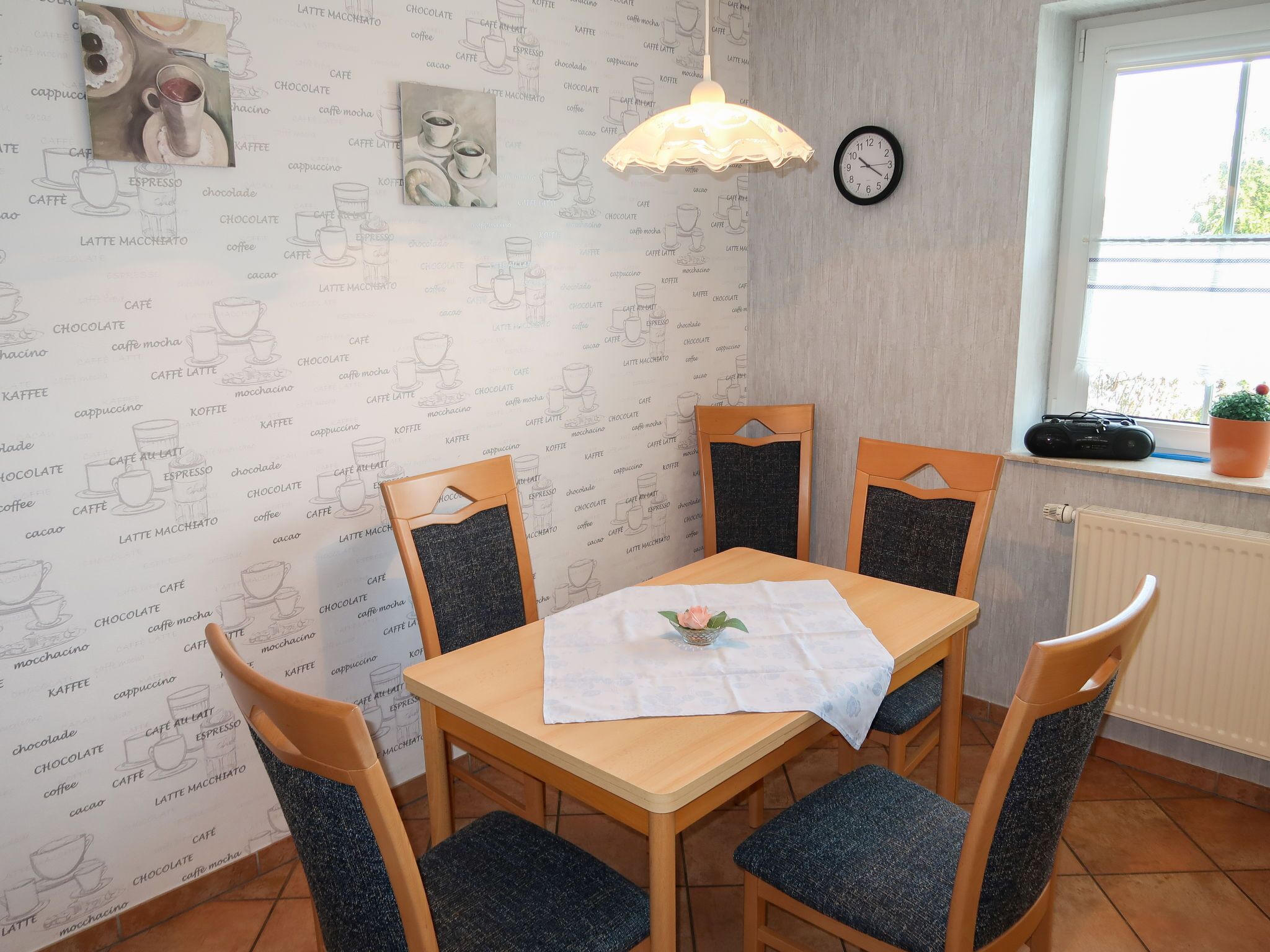A dining area with a wooden table, four chairs, a white tablecloth, a clock, and a window with a plant.