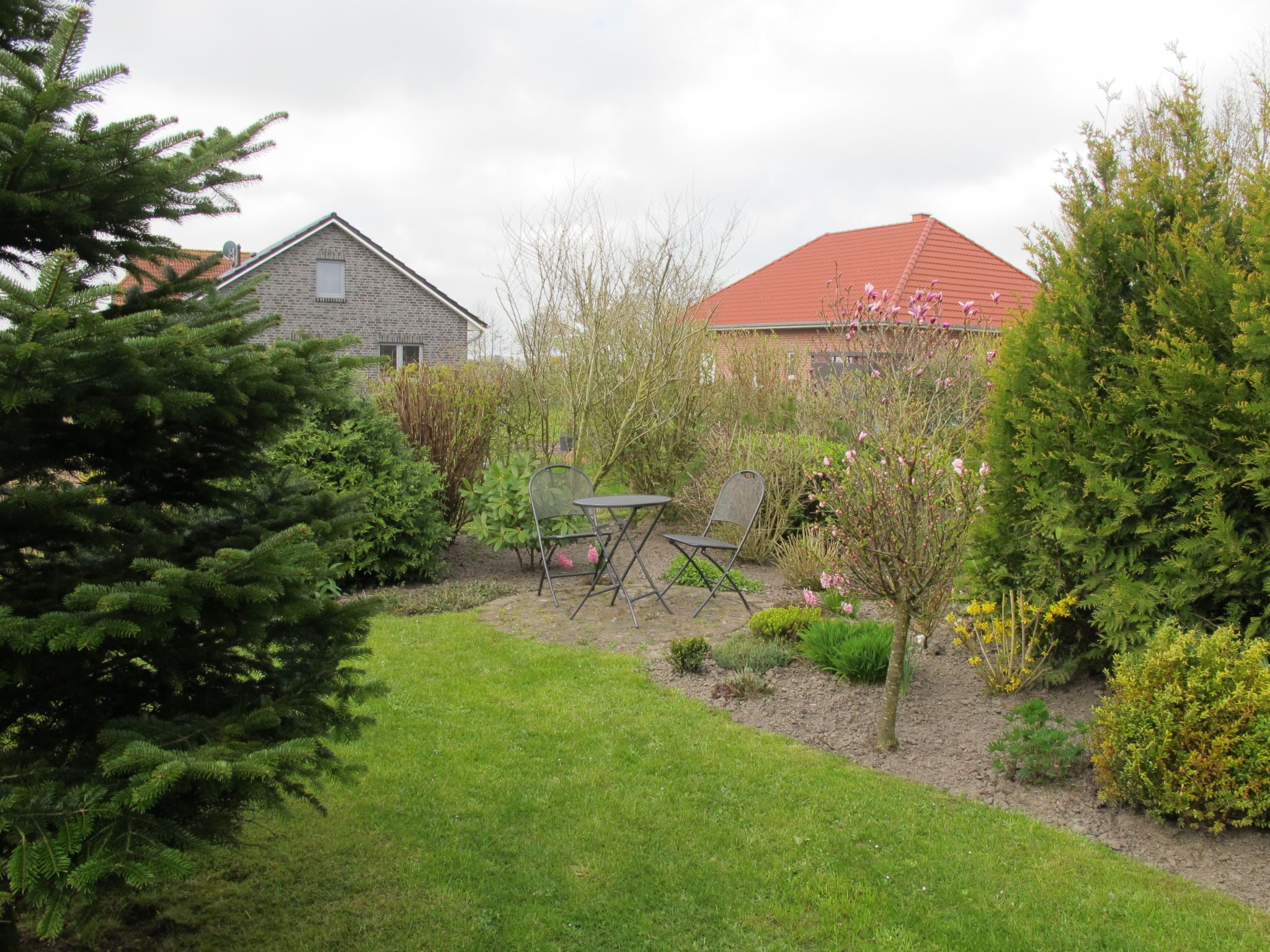 En trädgård med en gräsmatta, ett bord och några stolar, samt två hus med olika färg på taken.