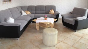 Un salon comprenant un canapé d’angle gris, un fauteuil noir, une table basse en bois et un pouf rond.