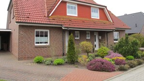 Une maison à deux étages, avec un toit recouvert de tuiles rouges, une façade en briques et un jardin devant, agrémenté de divers arbustes et fleurs.