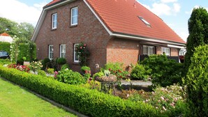Une maison à deux étages, construite en briques et surmontée d’un toit recouvert de tuiles rouges, est entourée d’un jardin bien entretenu où poussent différentes variétés de fleurs et d’arbustes.