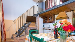 A dining area with a checkered tablecloth, green chairs, a fireplace, and a kitchen in the background. - Farfalla by Interhome (Riparbella)
