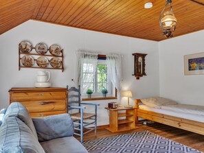 A cozy room with a wooden ceiling, a bed, a blue chair, a wooden dresser, a wall clock, and a window with curtains.