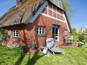 A red brick house with a thatched roof, a green door, and a small garden with a lawn chair and table.