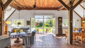 A cozy, open-plan living area with a wooden ceiling, a dining table, and a view of the garden through large glass doors.